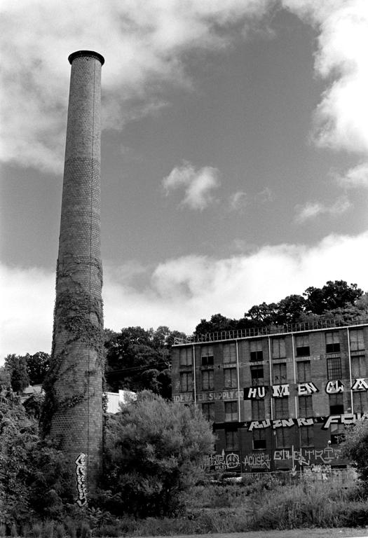 A tall brick chimney covered in ivy stands next to an abandoned building with graffiti under a cloudy sky.