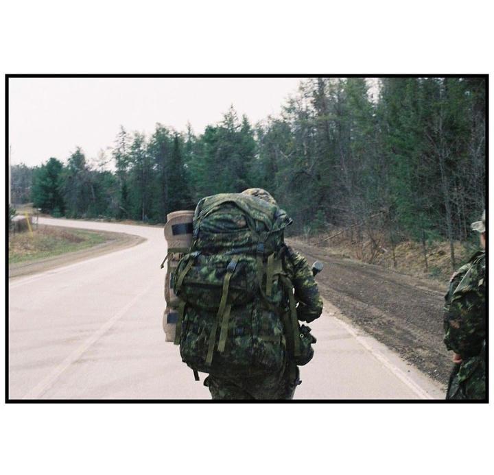 A person in camouflage with a large backpack walks along a road next to a forest.