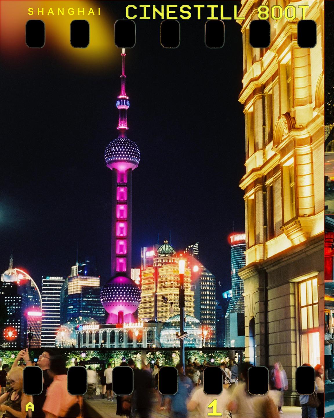 A crowd of people stands on a street at night with a tall, illuminated tower and other buildings in the background.