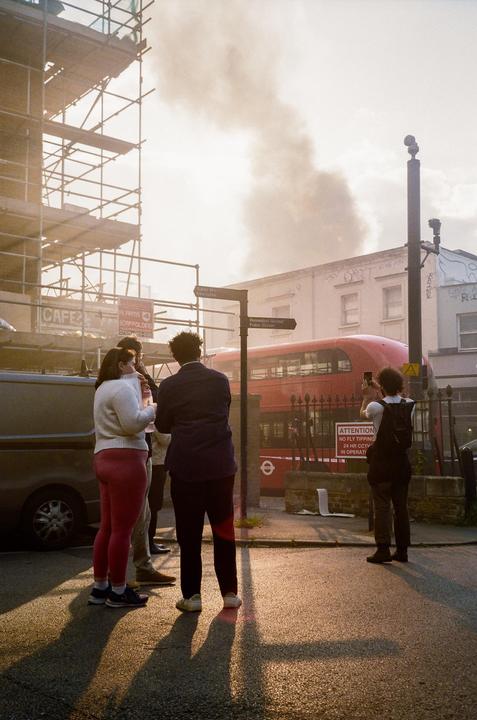 A group of people stand on a street corner watching smoke rise from a fire in the distance.