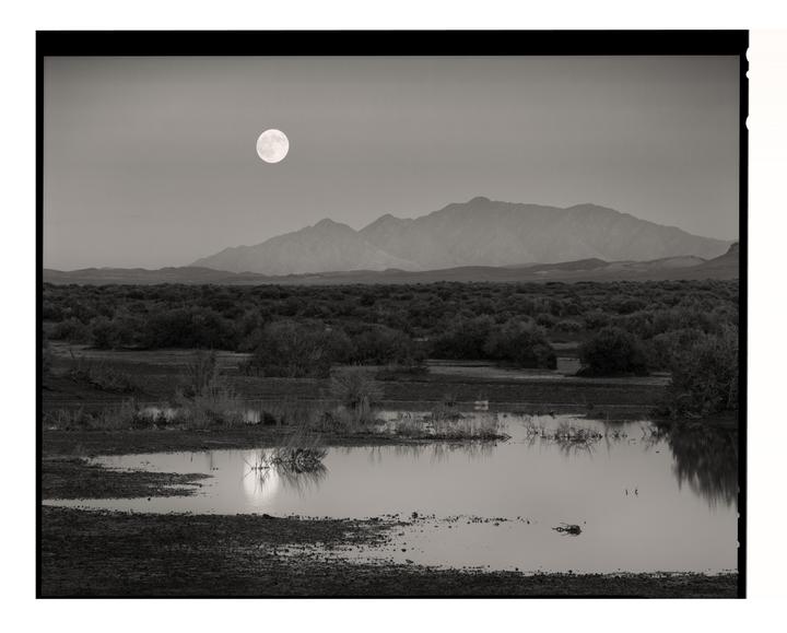 A full moon rises over a desert landscape with mountains in the distance and a body of water reflecting the moon in the foreground.