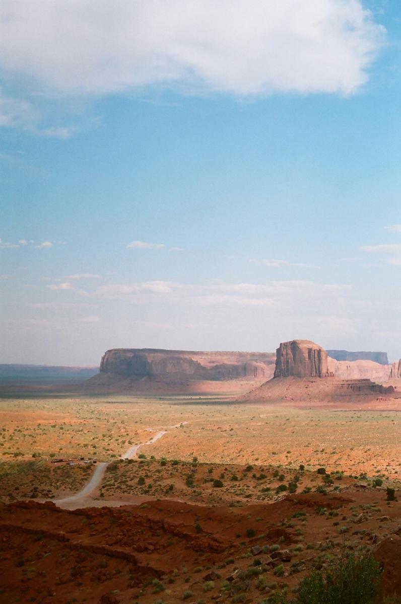 A dirt road leads through a vast desert landscape towards large rock formations under a blue sky with scattered clouds.