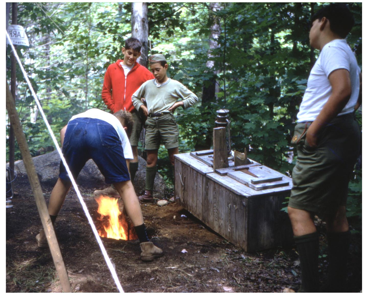 Four boys are gathered around a fire pit in a forest, with one boy tending to the fire.