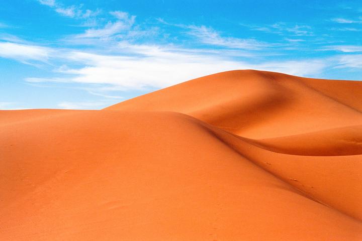 Rolling sand dunes are visible under a bright blue sky with wispy clouds.