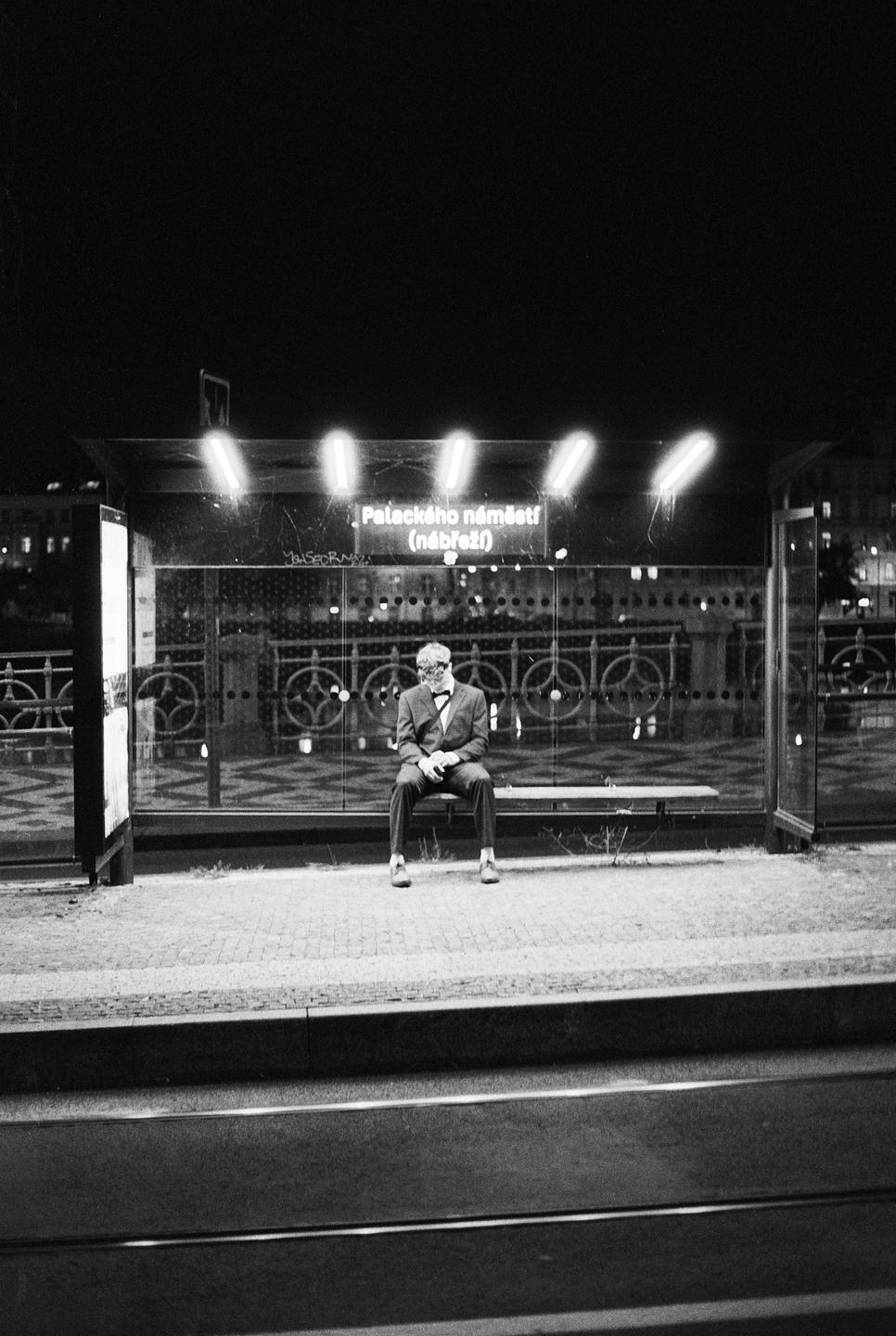 A man in a suit and bow tie sits on a bench at a bus stop at night.
