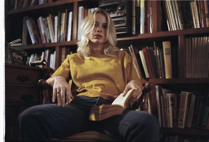 A woman with blonde hair sits in a chair, holding an open book, in front of a bookshelf filled with books.