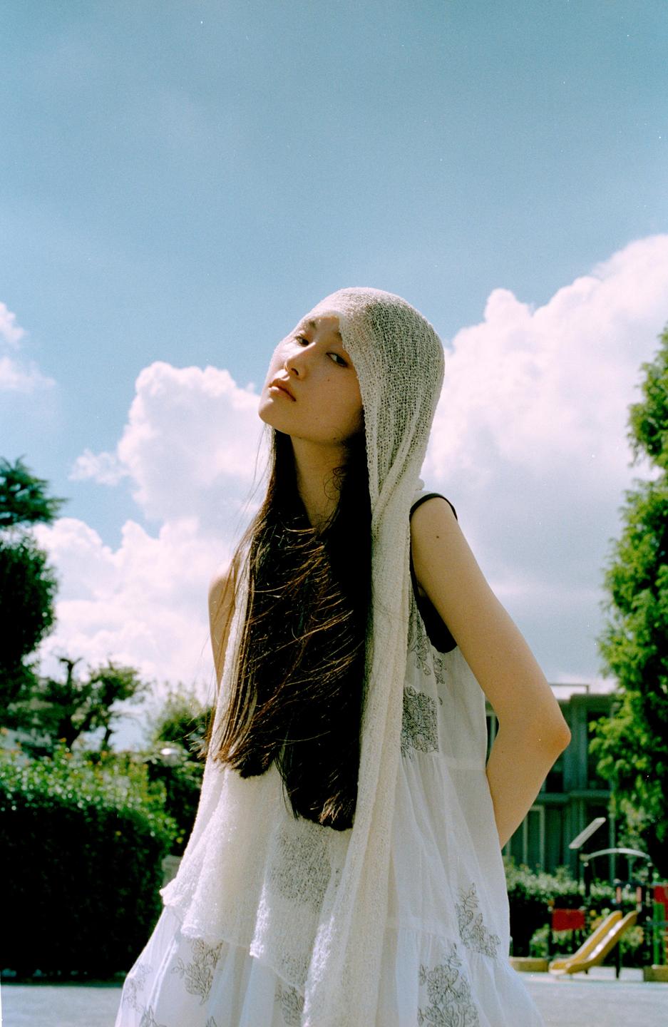 A woman with long dark hair and a white dress looks up at the sky with clouds in the background.