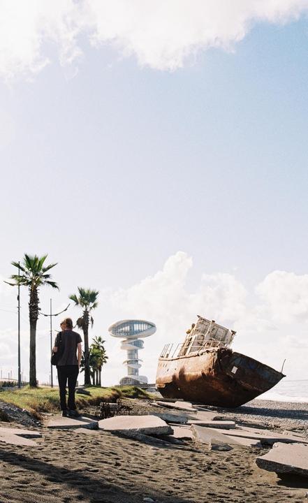 A man stands on a sandy beach with a rusted shipwreck and a modern building in the background.