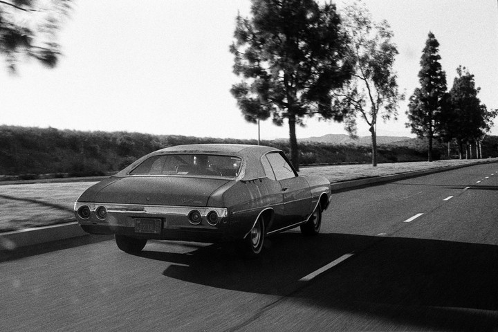 A classic car drives down a road lined with trees and hills in the distance.
