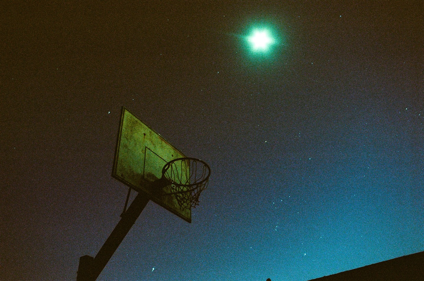 A basketball hoop stands against a starry night sky with a bright light source in the upper left.