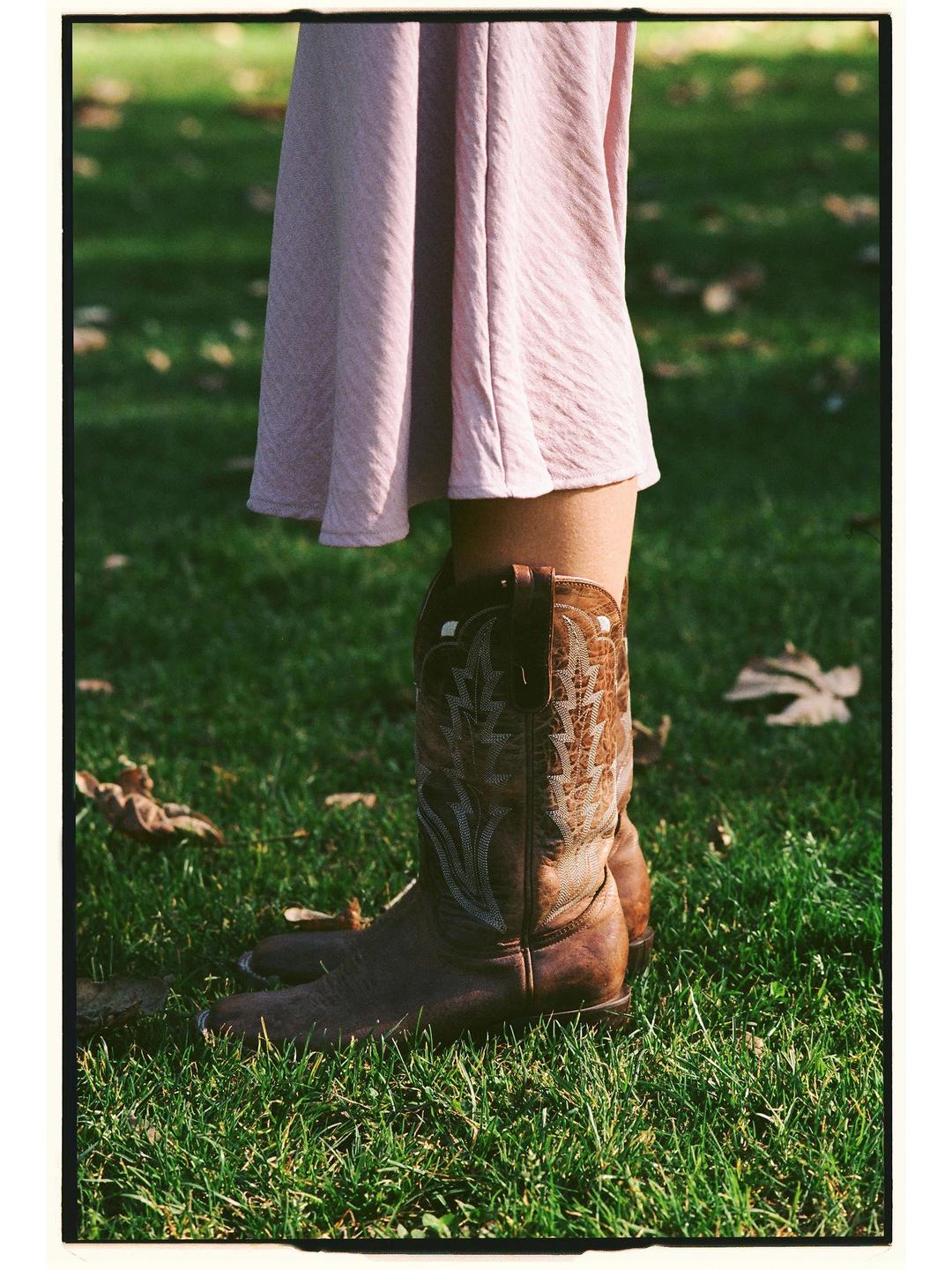 A person wearing a pink dress and cowboy boots stands in green grass with fallen leaves.