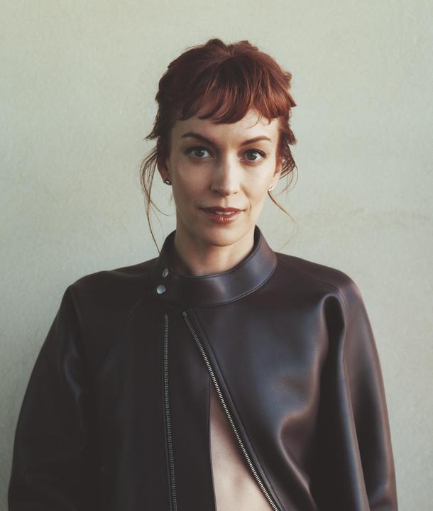 A woman with red hair and a brown leather jacket looks directly at the viewer.