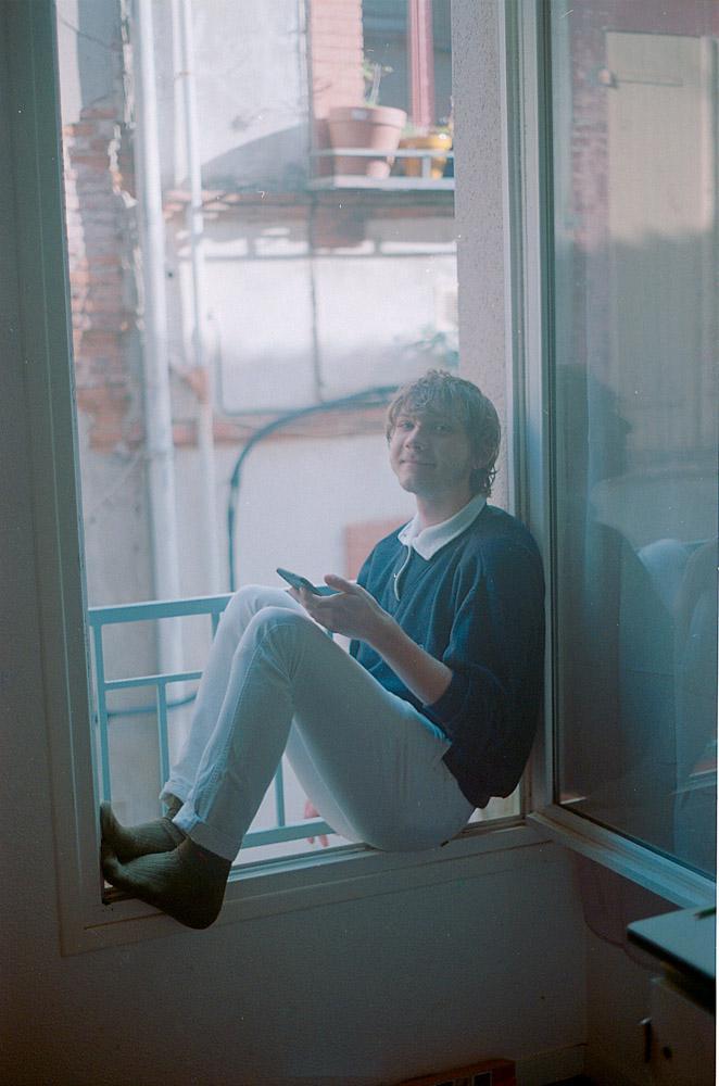 A man sits on a window ledge, holding a phone and looking out.