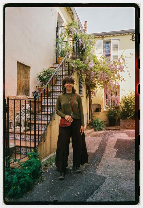 A woman stands on a paved path in front of a building with an outdoor staircase adorned with flowering vines.