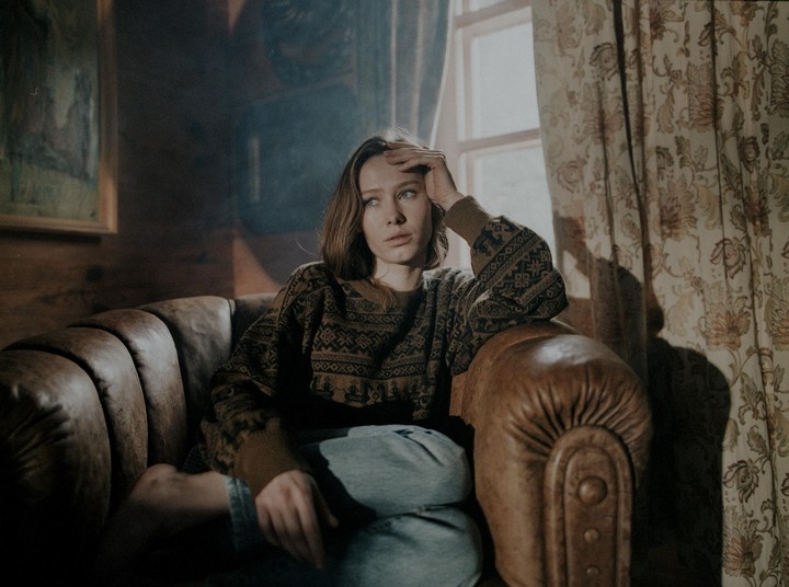 A woman sits on a leather sofa, looking away from the camera with her hand on her forehead, next to a window with patterned curtains.