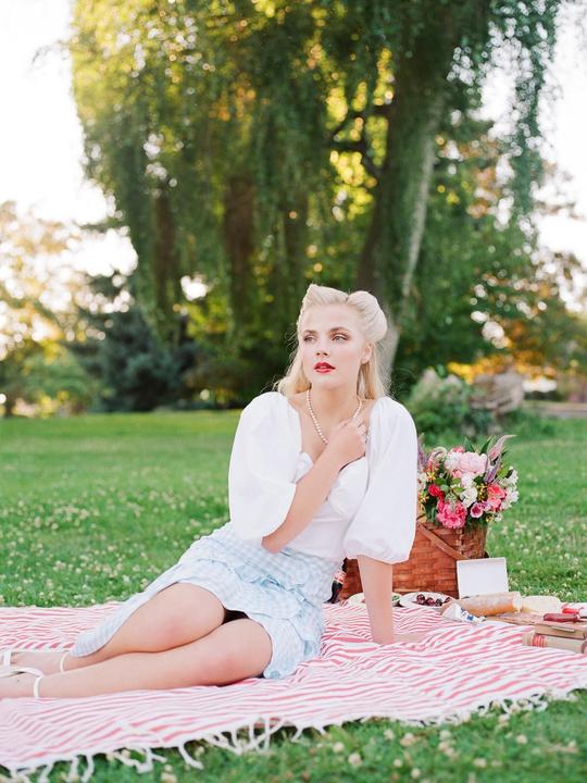 A woman in a white top and blue gingham skirt sits on a red and white striped blanket with a picnic basket and food next to her.