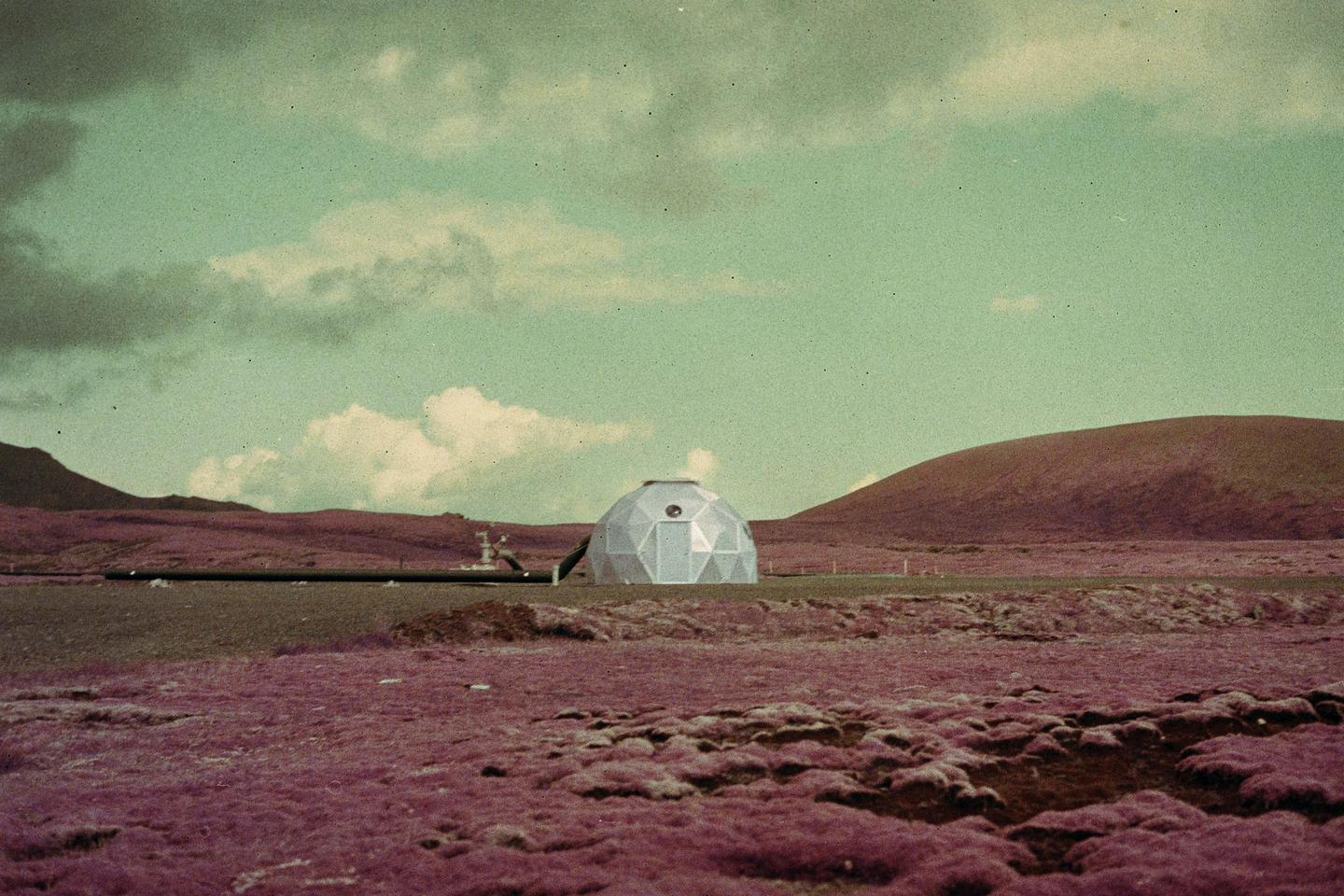 A geodesic dome sits in a purple field with hills in the background under a cloudy sky.