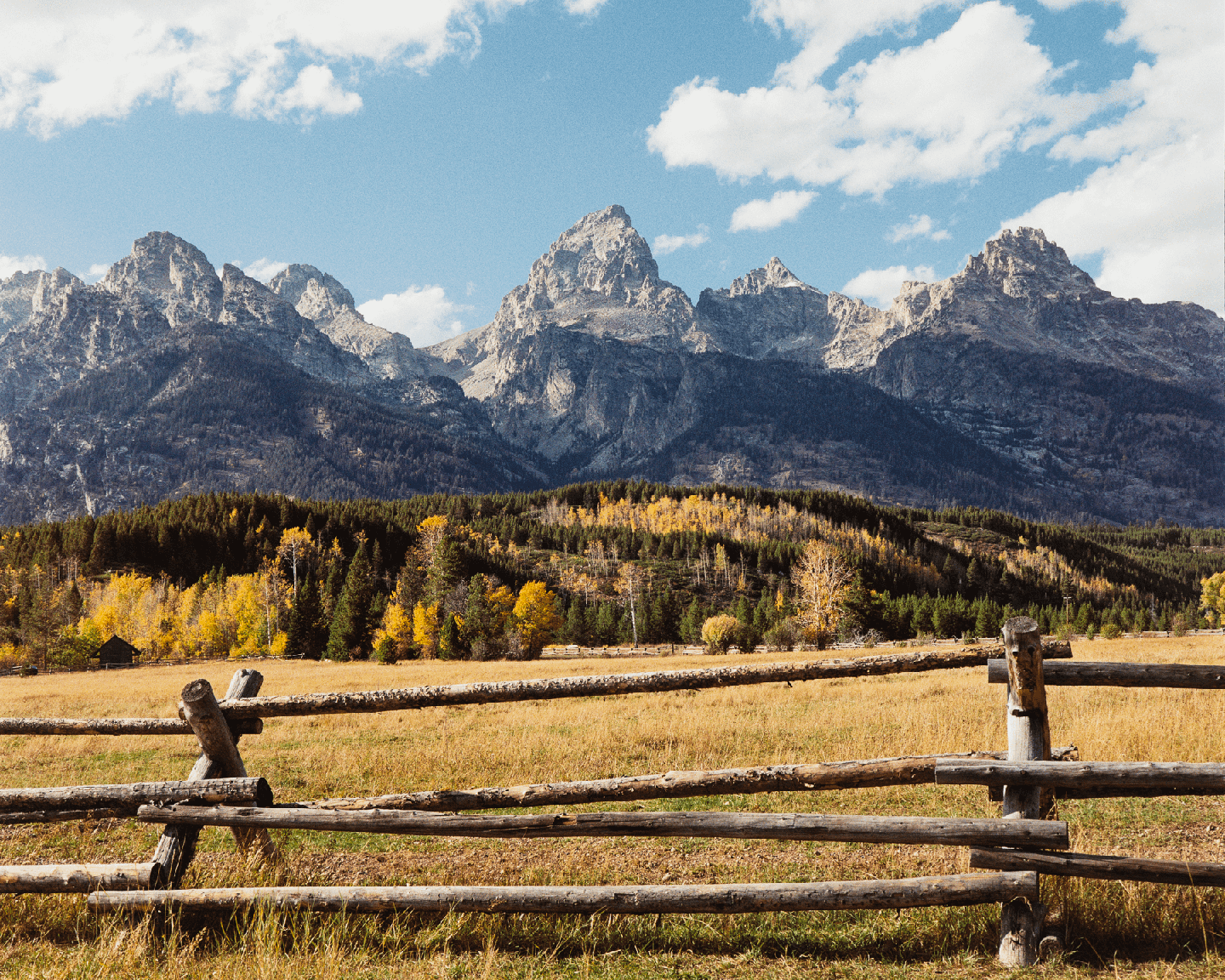 A wooden fence runs across a field with mountains and autumn trees in the background under a blue sky with clouds.