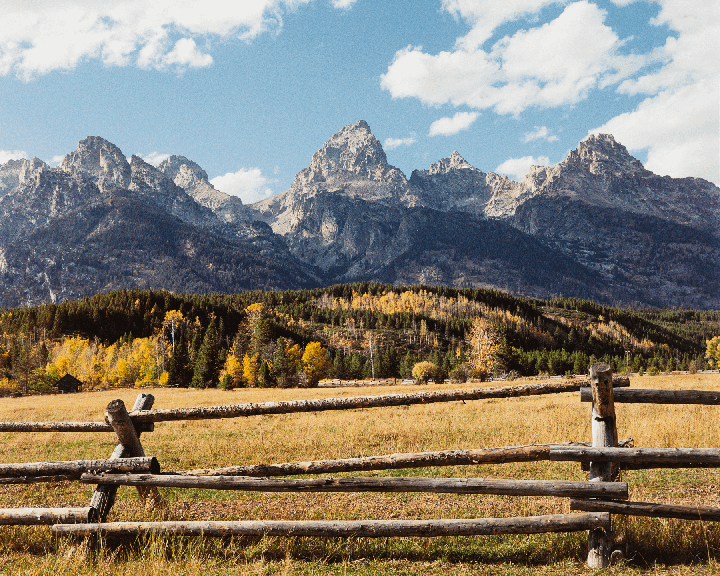 A wooden fence runs across a field with mountains and autumn trees in the background under a blue sky with clouds.