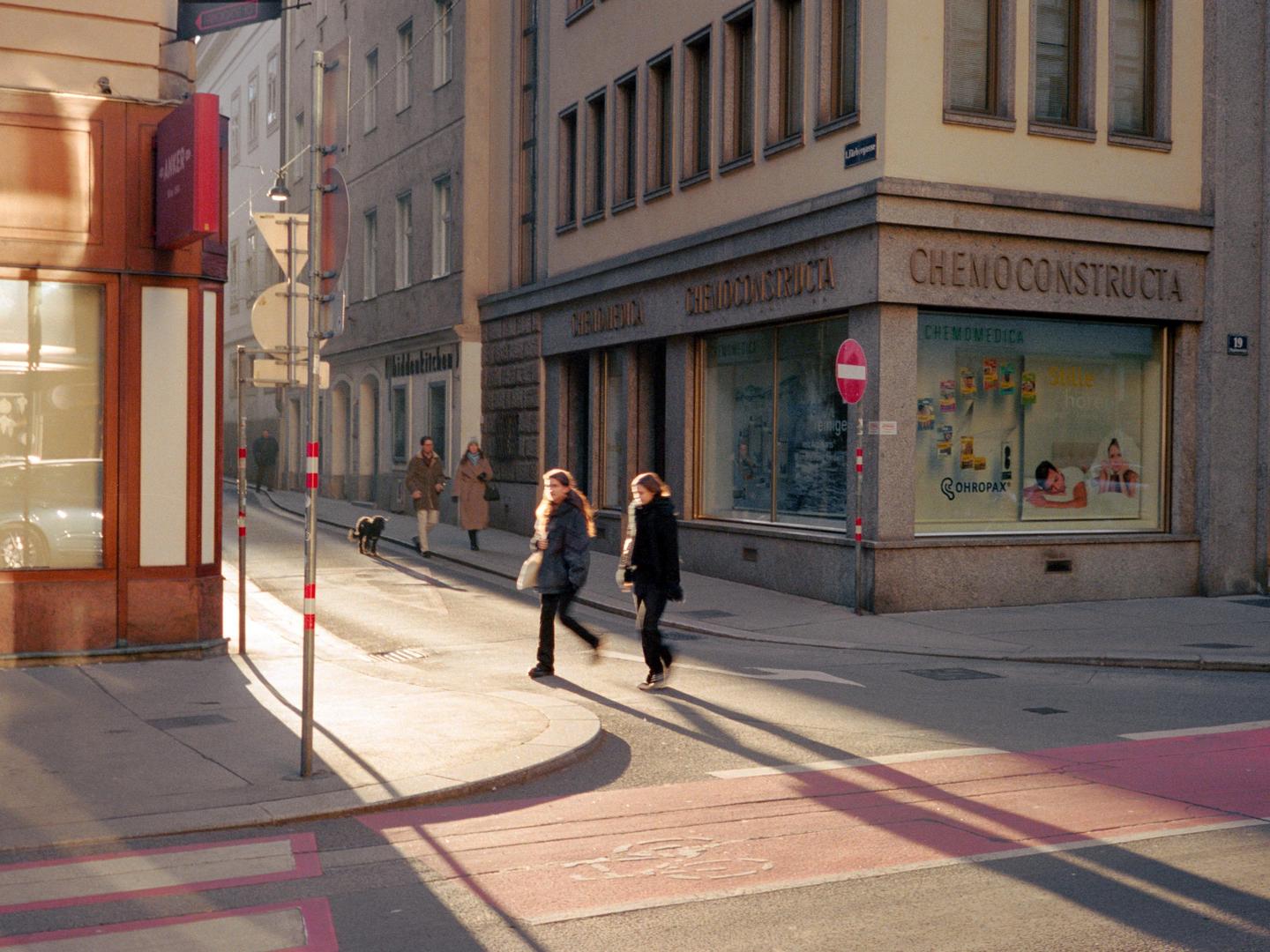 Two women walk across a sunlit street corner with long shadows stretching across the pavement.