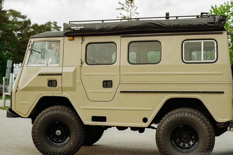 A man is visible through the driver's side window of a tan off-road vehicle with large tires and a roof rack.