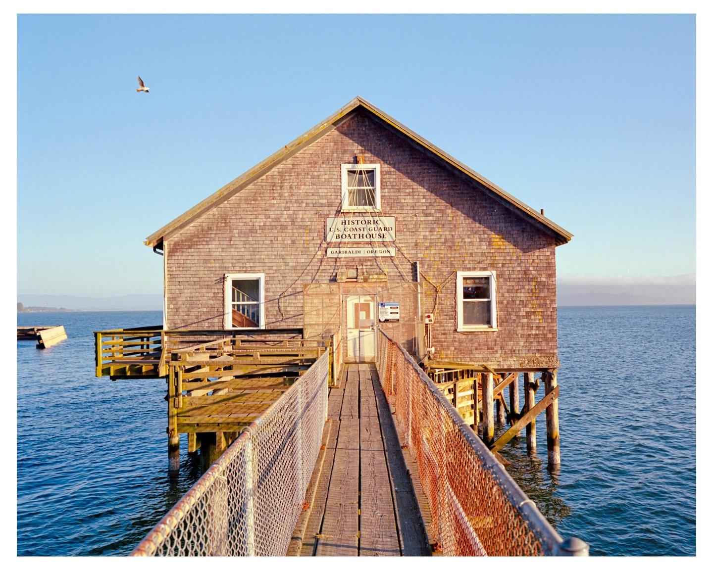 A wooden boathouse stands over blue water with a bird flying in the clear sky.