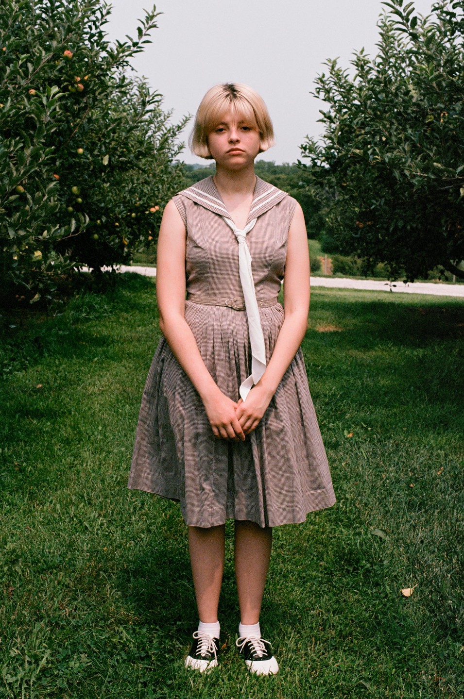 A young woman with blonde hair and a sailor-style dress stands in a grassy field between two apple trees.