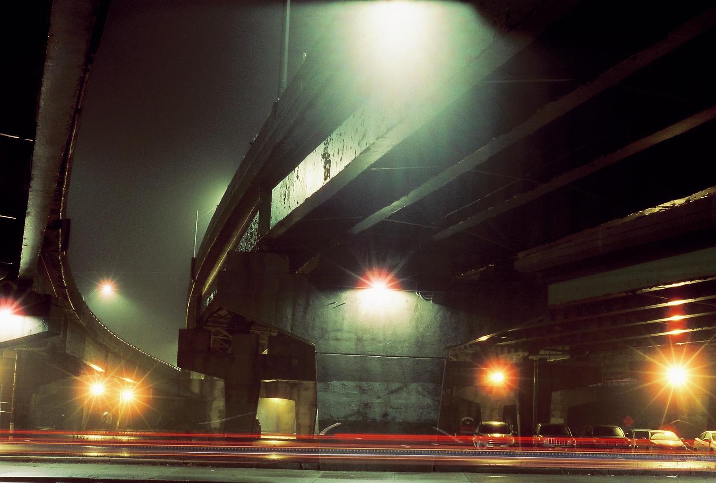 A long exposure shot captures the light trails of vehicles under a highway overpass at night.
