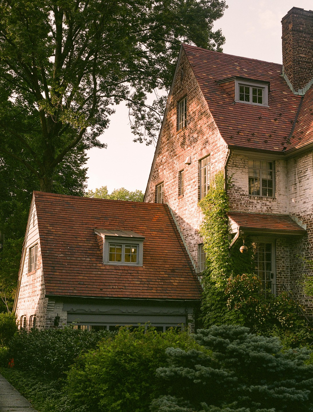 A large brick house with a red tile roof is surrounded by lush green trees and bushes.
