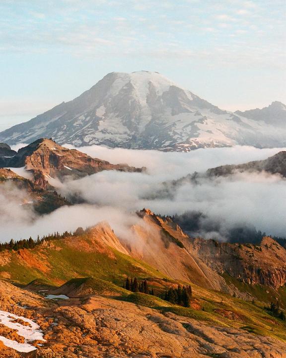 A snow-capped mountain rises above a layer of clouds, with sunlit hills and trees in the foreground.
