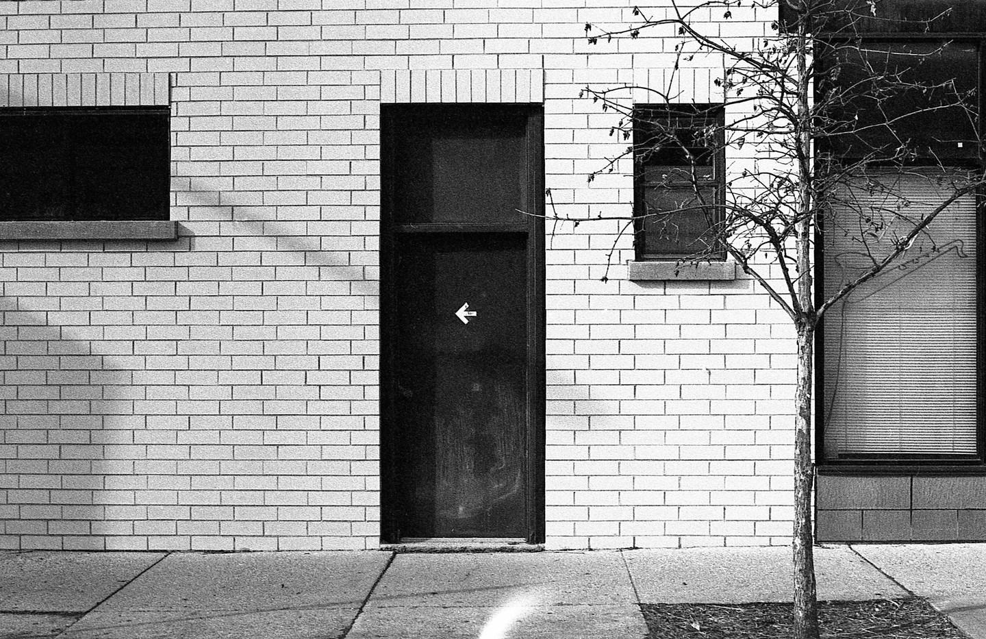 A black door with a white arrow pointing left is set in a white brick wall next to a bare tree and windows.