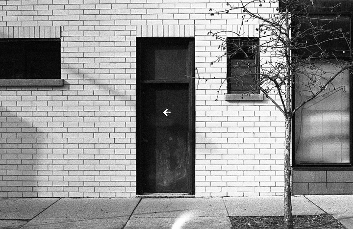 A black door with a white arrow pointing left is set in a white brick wall next to a bare tree and windows.