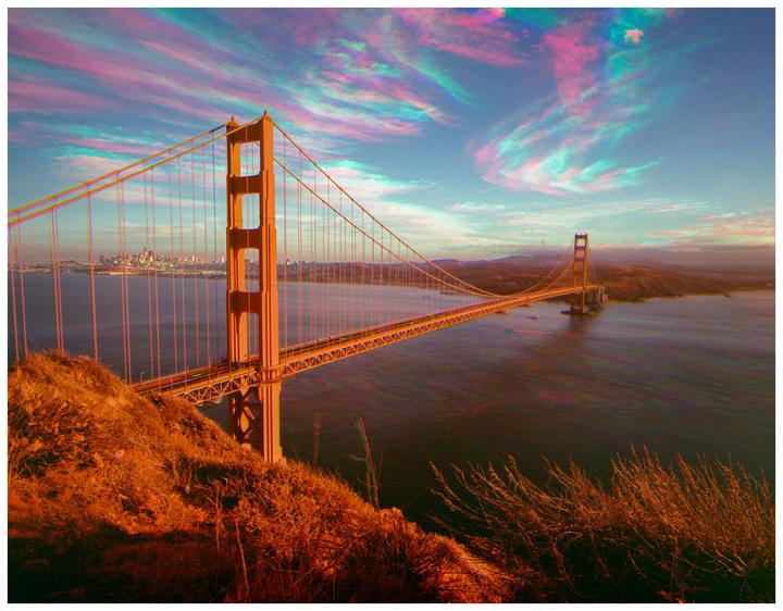 The Golden Gate Bridge spans a body of water with a city skyline in the distance under a sky with colorful clouds.