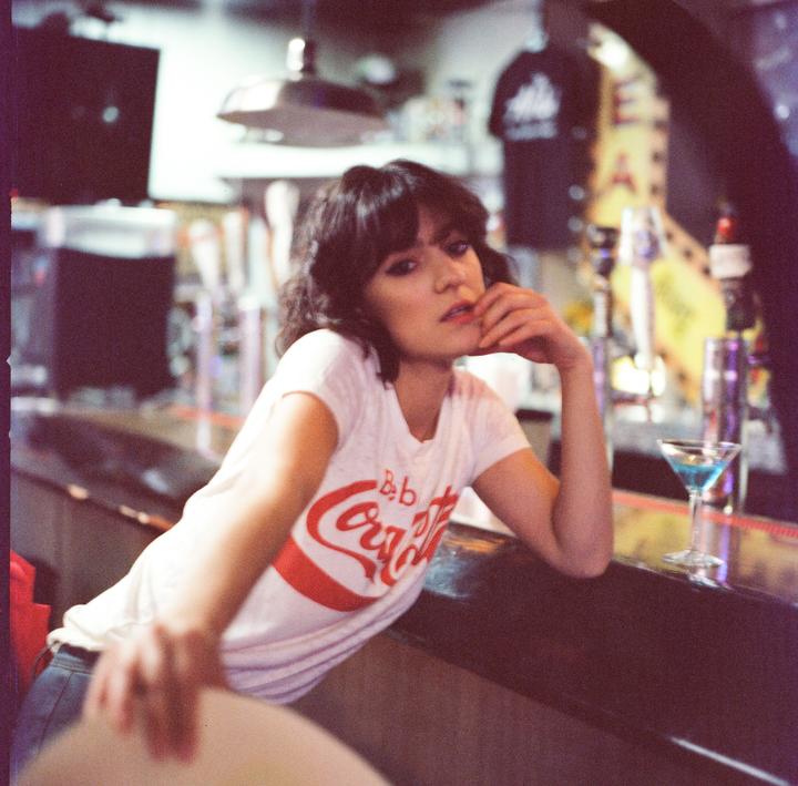 A woman in a white t-shirt with a red logo leans on a bar next to a blue cocktail.