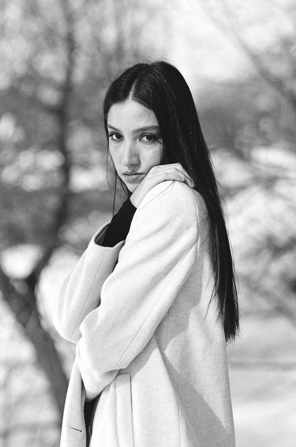 A woman with long dark hair wears a light-colored coat and looks directly at the viewer.