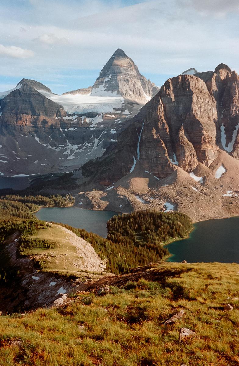 A mountain range with snow-capped peaks overlooks a valley with two lakes and a dense forest.