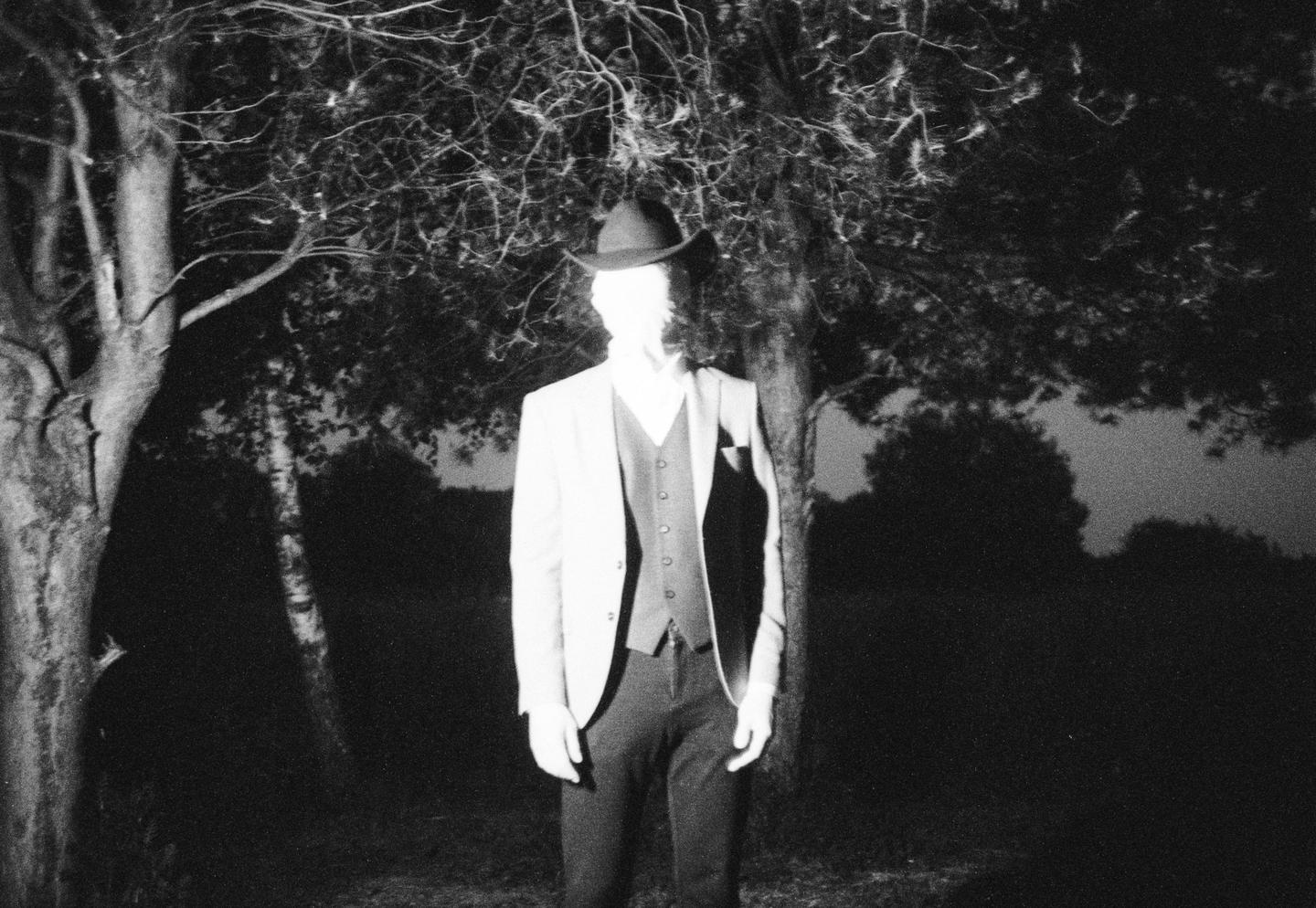A man in a suit and hat stands in a forest at night.