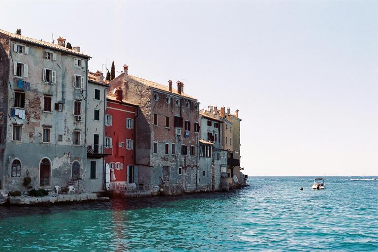 A row of colorful buildings with shuttered windows stands along the edge of a bright blue sea with a small boat.