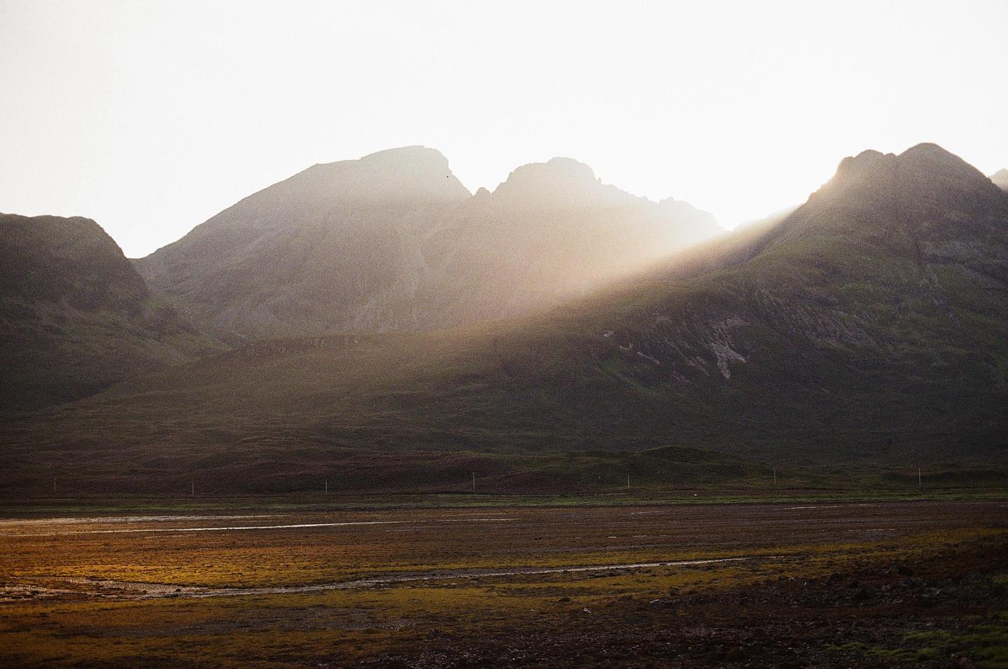 Sunlight streams over a mountain range and illuminates a field in the foreground.