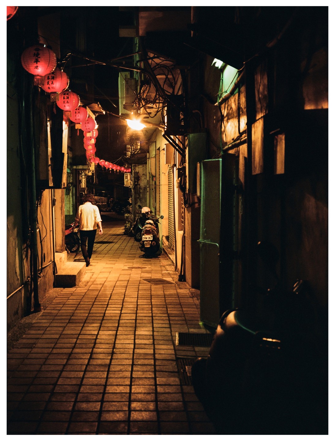 A person walks down a narrow alleyway at night, illuminated by red lanterns and streetlights.