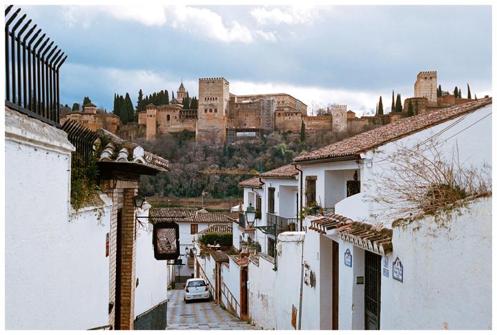 A narrow street with white buildings and terracotta roofs leads down to a small white car, with a large castle on a hill in the background.
