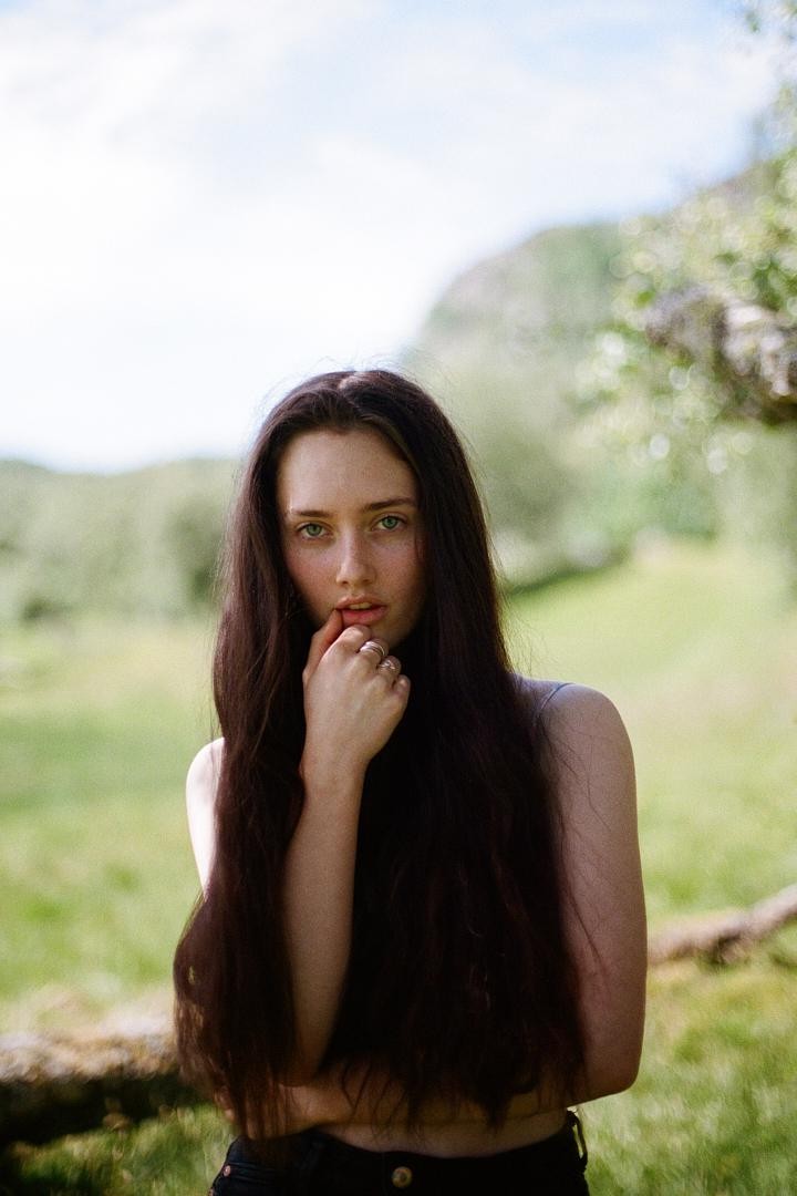 A woman with long dark hair looks directly at the viewer with her hand near her mouth in a grassy outdoor setting.