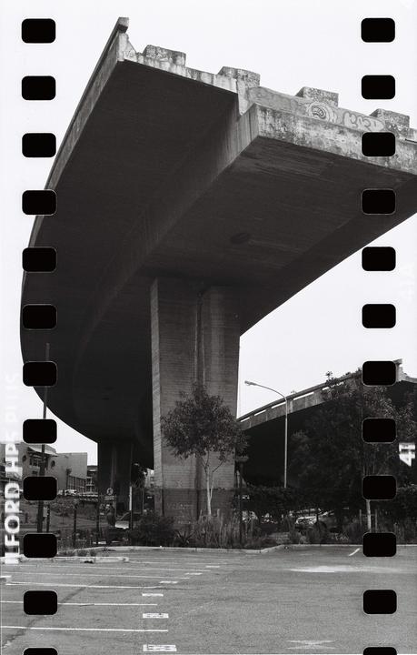 A large concrete overpass with graffiti on its underside looms over a parking lot and trees.