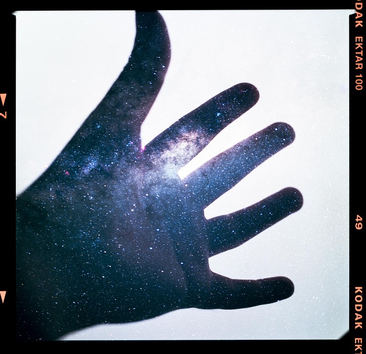 A hand is silhouetted against a bright background, with the milky way visible through the hand.