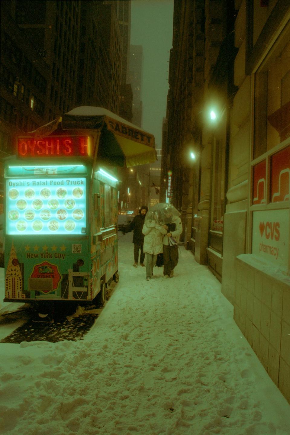 A halal food truck with glowing signs is parked on a snow-covered street in a city at night, with people walking past.