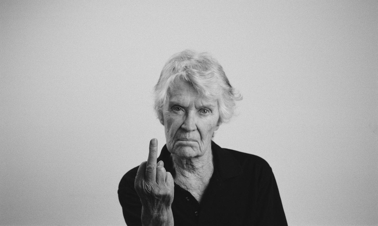 A woman with short, wavy hair and a black shirt gives the middle finger to the camera.