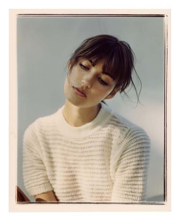 A woman with dark hair and bangs wears a cream-colored sweater and looks down.
