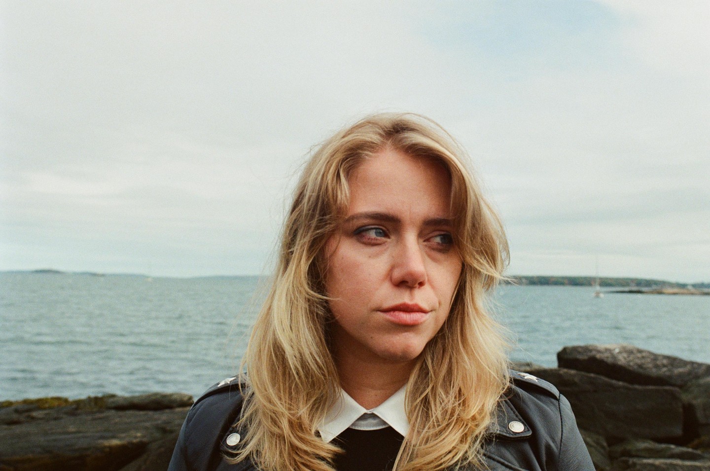 A woman with blonde hair and a leather jacket looks away from the camera with the ocean and a cloudy sky behind her.