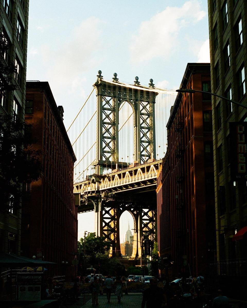 The Manhattan Bridge is framed by two buildings and a street with people and cars.