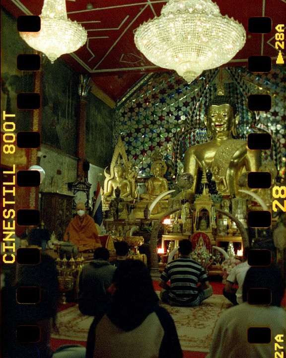 People sit on the floor of a temple with golden buddha statues and two large chandeliers.